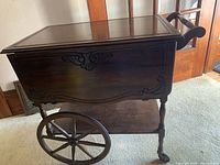 View of side of the tea cart showing one drop leaf down, large wooden wheel with rubber, decorative wood carvings, and turned legs.