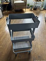 Full view of the gray metal rolling shelf with three mesh trays on caster wheels, shown indoors on wooden floor.