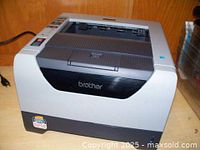 Photo of a Brother laser printer on a wooden surface showing front and top with paper tray and control buttons.