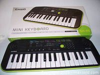 Casio Mini Keyboard SA-46 with its original box showing the front of the keyboard with 32 keys and multiple control buttons, including a green dial, on top. The keyboard is black with white and black keys, green accents along the edges.