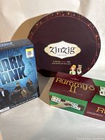 Photo showing three board games: Shark Tank the Game in blue box, Zinziz round wine tasting game box, and Rummy-O II tile game box.