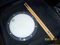 Photo shows a Remo drum practice pad with a white playing surface and black rim with metal tension bolts, resting on a black textured surface alongside a pair of natural wood drumsticks.