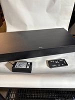 Photo showing black rectangular Zvox sound bar with power adapter and remote control on white backdrop