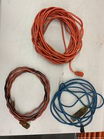Three coiled extension cables laying on a white surface: two are orange and one is blue. All have standard plug and socket ends.