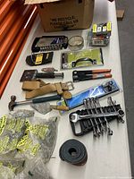 Various hand tools and hardware including hammer, wrenches, screwdriver, wire cutters, screw bags, belt, and putty knife laid out on a surface.