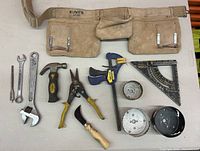 Photo of tools including Kuny's tool belt, crescent wrench, open-end wrenches, hammer, locking pliers, clamps, square protractor, tape measure, pruner, and hole saw blades