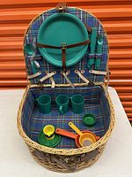 Open picnic basket showing turquoise plates, cups, spoons, knives, forks, and colored measuring spoons with small plates inside on tartan fabric lining.