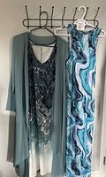 Two dresses hanging on a wall rack: one light blue sheer overlay with a paisley patterned dress, and a long sleeveless dress with marbled swirl print and large back cutout.