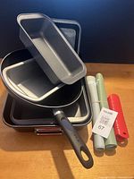 Stacked set of non-stick bakeware pans including a springform pan with black handle latch, two rectangular pans and a red silicone handle grip alongside two rolled silicone baking mats with leaf motifs.