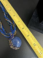Close-up showing moon style pendant with detailed blue stone and peacock feather gold/blue adornment next to measuring tape showing pendant approx 2.5 inches.