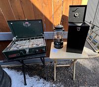 Overall photo showing 2 Coleman camping items (stove and lantern) placed on 2 wooden folding tables outdoors on gravel with snow patches.