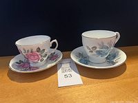 Pair of vintage teacups with matching saucers on wooden surface, one set with pink roses and one set with blue roses, both with gold trim.