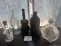 6 collectible bottles and decanters displayed in front of lace curtain including Kinsip bottle, violin shaped bottle, and clear cut glass decanter.