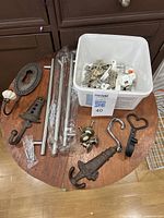 Various hooks, handles, hinges, and hardware pieces arranged on a round wooden table. Includes dark bronze decorative hooks, tubular silver handles, and plastic container with small hinges and screws.
