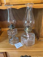 Two glass hurricane lamps side by side on shelf, showing bases and chimneys with metal burners.