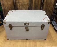 Front closed view of large grey metal chest with brass cornering, clasps, and leather handles, showing scratches on paint.