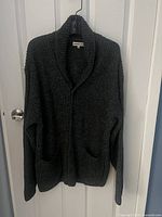 Full view of black London Fog cardigan with two front pockets, shawl collar, hanging on a door.