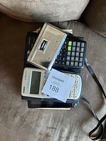 Top view photo showing two calculators, a metallic item (likely binoculars), and other electronics stacked together.