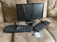 Monitor with two keyboards and a mouse on a brown couch. One keyboard has PS/2 connector and shows vintage style and wear; the other keyboard and a mouse with USB connectors appear modern.