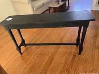Wooden black plant stand displayed in a living room setting on a hardwood floor, showing front and side view with turned legs and stretcher.