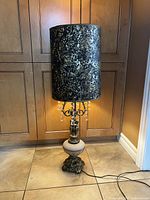 Full lamp view showing tall brass lamp with ornate glass base and cylindrical black and gold lampshade lit from within.