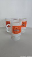 Three vintage white pedestal coffee mugs showing the front side with The Toronto Sun circular logo inside an orange square.