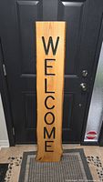 Full view of the tall wooden sign leaning against a black door, showing the entire 'WELCOME' text vertically from top to bottom.