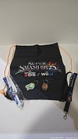 Full group of Nintendo promotional items: black Super Smash Bros drawstring bag, two character keychains, Nintendo wristband, and lanyard displayed on white surface.