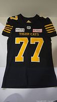Full front view of black Hamilton Tiger-Cats jersey featuring yellow and white number 77, sponsor patches, and embroidered team name.