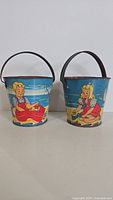 Front view showing both pails side by side with printed beach scenes of children and dog on blue backgrounds.