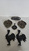 Seven vintage cast iron and brass trivets and decorative items arranged on white background, including two rooster trivets, two brass face motifs, small sailboat, and two cast iron animal oval trivets.