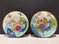 Front view of two circular decorative bowls with colorful floral and tobacco leaf designs on porcelain centers encased in brass.
