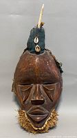 Front view of the African wooden tribal mask showing geometric facial carving, fabric horn with cowrie shells, and fiber beard.