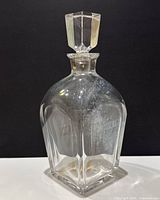 Clear glass decanter with faceted square stopper standing upright on black background showing overall shape and condition.