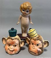 Front view of Kewpie doll with jointed arms on wooden stand alongside two Shafford monkey head salt and pepper shakers with colorful tops.