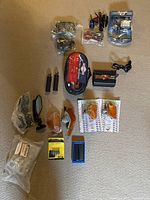 Lot items arranged on carpet including booster cables, mirrors, foot pegs, orange tail light covers, power connectors, rear view camera display, riser set, solar charger, power inverter.