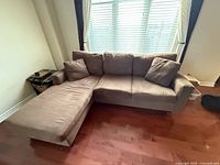 Photo of brown fabric sofa with chaise on left side, three seat cushions, three back cushions, two matching pillows, situated on wooden floor against window with blinds.