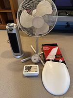 Photo showing silver electric heater, white pedestal fan, white plastic toilet seat, first aid kit, and smoke and carbon monoxide detector placed on carpeted floor in front of wooden furniture.