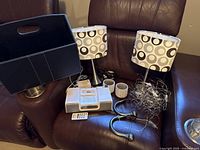 Photo of the entire lot including two brushed silver lamps with patterned shades, brown leather magazine/book holder, tangled USB fairy lights, iHome clock radio with remote, and two small clay flower pots.