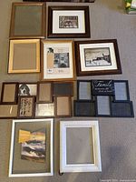 Top view of thirteen assorted picture frames laid out on a beige carpet; various frame styles, colors, and sizes; many frames without pictures, some with placeholder prints or photographs.