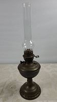 Full view of the copper oil lamp with tall clear glass chimney showing overall shape and condition.