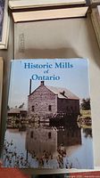 Photograph of the front cover of the Historic Mills of Ontario book with a stone mill building and pond reflection on dust jacket
