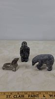 Three Inuit soapstone carvings on a light background: a hunter figure in black with hand damage, a composite polar bear, and a seal figure.
