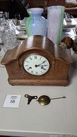 Front view of oak mantle clock on table with two glass vases behind, brass pendulum and winding key placed in front