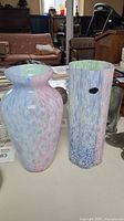 Photo showing two pastel-colored glass vases, one wide curved and one tall cylindrical on a table.