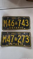 Front faces of two black Ontario license plates from December 1975 with yellow embossed characters and age wear.