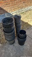Multiple stacks of large, medium, and small black plastic potting containers arranged outdoors on concrete next to a brick wall.