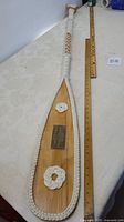 Full view of the 60-inch wooden presentation paddle with rope decorations and commemorative plaque, shown next to ruler for size.