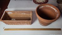 Two terracotta pots side by side with ruler showing scale. Rectangular planter on left, round flowerpot on right.