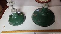 Two green enamel pendant lights side by side on a white surface, one is 12 inches wide and the other is 16 inches wide showing worn areas